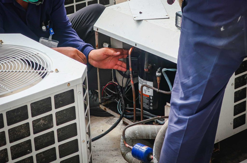 Technicians in blue uniforms repair an HVAC unit, surrounded by open electrical panels and wires, conveying focus and teamwork on a rooftop.